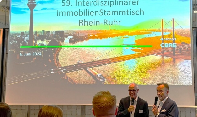 A screen displays the title of the event; two people with microphones stand in front of it on the stage.