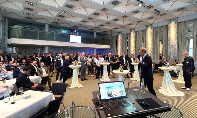 Event hall, view from the stage of many people, some standing at tables.